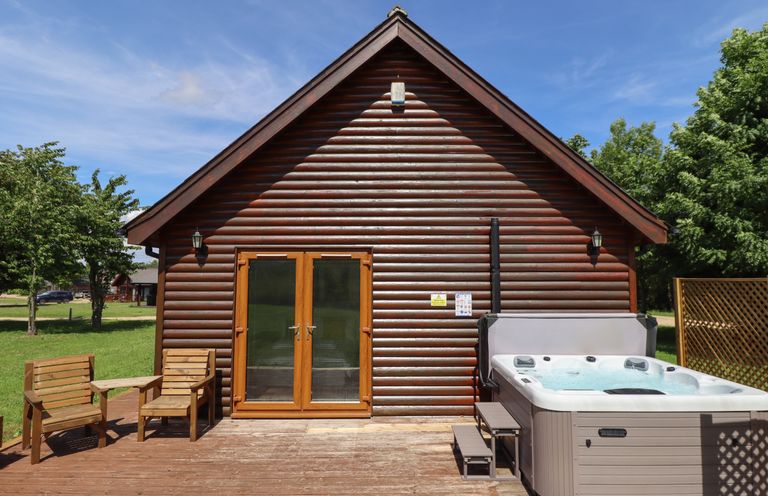A wooden cabin with glass doors a hot tub and two wooden chairs on a deck at Lodge 1 in South Hykeham