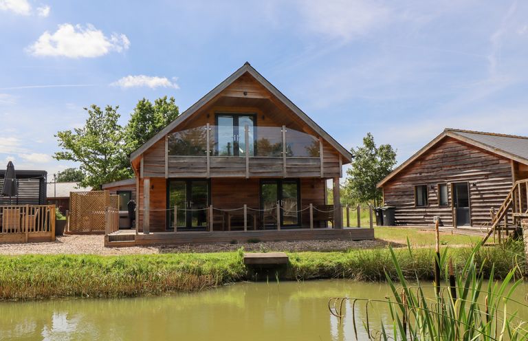 A wooden lodge with balcony and glass doors next to a pond and grassy area at Lodge 24 in South Hykeham