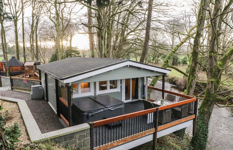 A cabin with a covered hot tub and outdoor seating on a deck overlooking a river surrounded by trees at Buttermilk Bridge in Dungiven