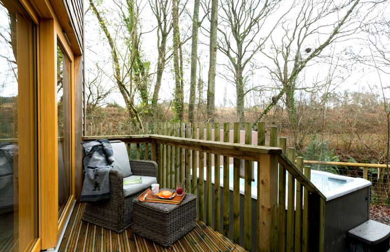 A wooden balcony with a wicker chair and table next to a hot tub surrounded by trees at Lodge 13 in Corfe Castle