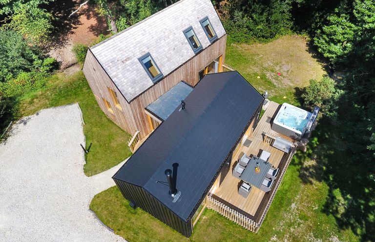 Aerial view of a two section wooden house with a deck that has a dining table with chairs and a hot tub surrounded by grass and trees at Lodge 20 in Corfe Castle