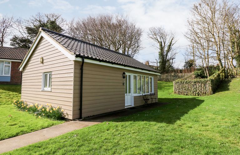 A small beige house with a tiled roof on a grassy lawn with a pathway and wooden fence at 12 The Dell in Mundesley
