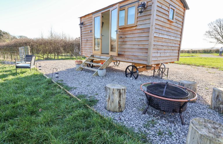 A wooden shepherds hut on wheels with open doors beside a fire pit and log stools at Cariad Bach in Gwaenysgor near Prestatyn