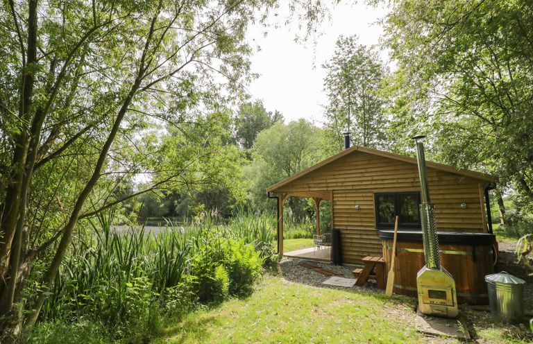 A wooden cabin with a covered porch and outdoor wood stove next to a grassy area and trees at Kingfisher in Dunchurch