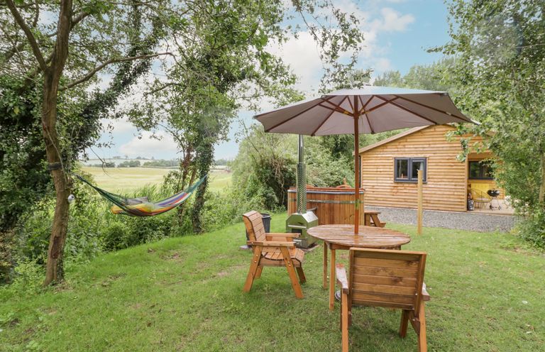 An outdoor garden area with wooden chairs a table with umbrella a hammock trees and a wooden cabin at Dragonfly in Dunchurch