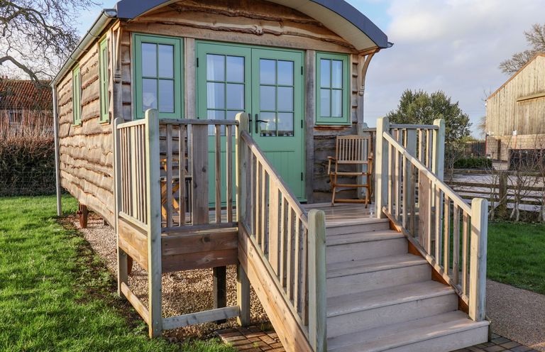 A wooden cabin with green doors and windows a porch with stairs and wooden chairs at The Studio in Bolton Percy near Tadcaster