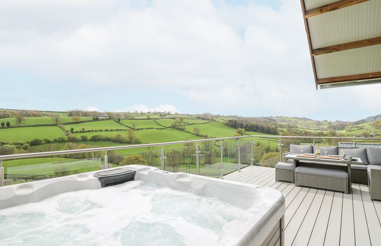 An outdoor deck with a hot tub glass railing and outdoor seating overlooking green hills at Barcud Coch - Red Kite in Garthmyl