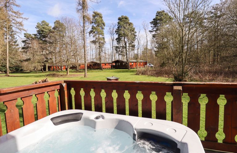 A hot tub on a wooden deck overlooking a grassy area with trees and wooden cabins in the background at iLodge 73 Kenwick near Louth