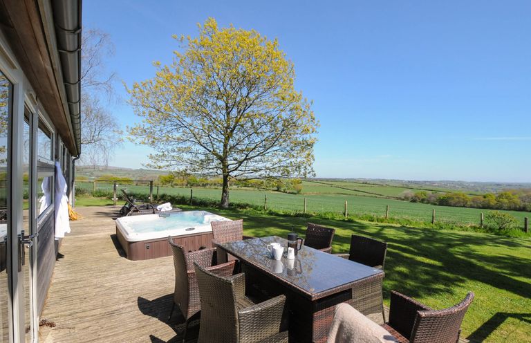 An outdoor wooden deck with a glass table and wicker chairs near a hot tub and a tree with fields in the background at 10 Faraway Fields in Dobwalls