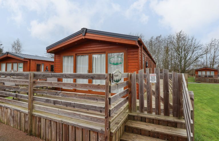 A wooden cabin with a fenced deck and stairs on a grassy area at Pegasus in Felmoor Park
