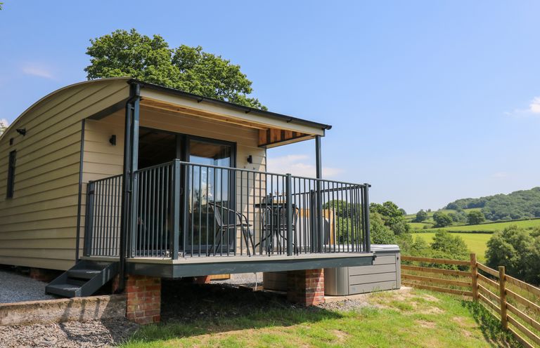 A small raised cabin with a balcony and outdoor chairs overlooking a grassy landscape at Rooster's Rest in Rhaedr Gwy near Rhayader