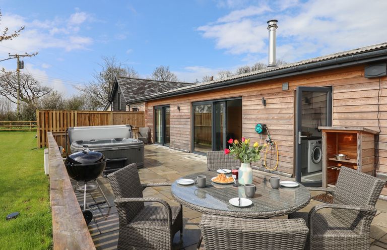 An outdoor patio area with a round glass table and wicker chairs a barbecue grill a hot tub and a wooden building in the background at Strawberry Bank - Nuthatch in Pyworthy near Bridgerule