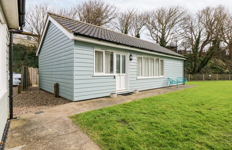 A single-story house with blue exterior siding white door and windows two blue chairs on the patio and a grassy lawn at 1 The Dell in Mundesley