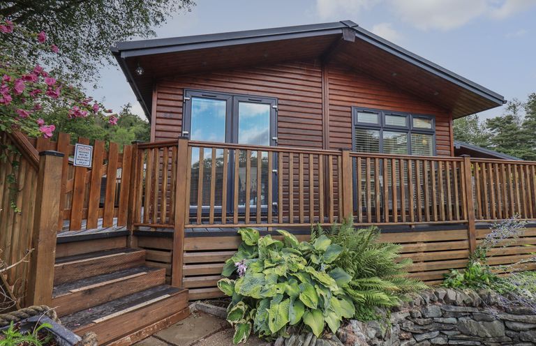 A wooden cabin with a deck and railing surrounded by plants at Hill Top Lodge in Bowness 65 White Cross Bay