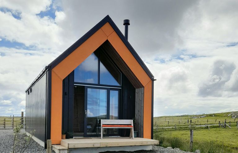 A small modern cabin with orange and black accents and a bench on the porch in a grassy rural area at North Light Studio in Locheport near Lochmaddy North Uist