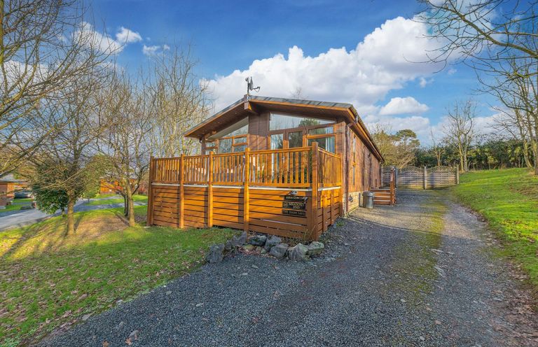 A wooden cabin with a fenced deck and gravel driveway surrounded by trees at Fern Lodge in Allithwaite near Cartmel