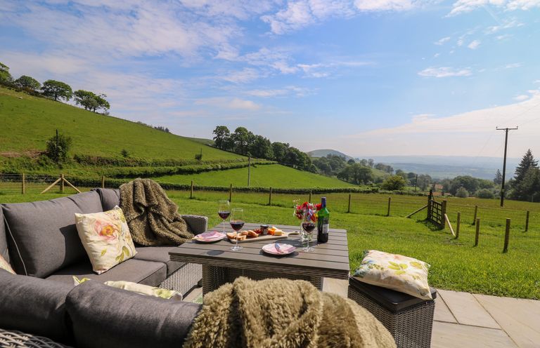 An outdoor seating area with gray cushioned chairs pillows a table with wine glasses and snacks overlooking green fields at Dol Blodau in Newbridge-On-Wye