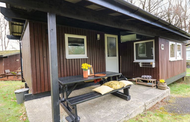 A black picnic table with pillows and a flower pot on the porch of a wooden cabin at 64 Penlan Holiday Park Cenarth