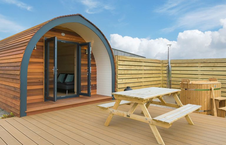 A wooden pod with open doors next to a picnic table and a wooden hot tub on a deck at Sun Rise in St Buryan