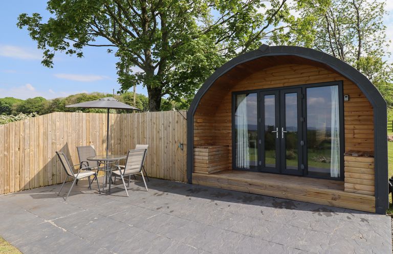 An outdoor patio area with a table and four chairs under an umbrella next to a curved wooden cabin at Dryfe Lodge in Lockerbie