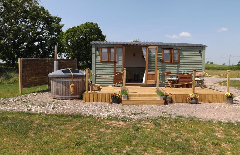 An outdoor wooden deck with a seating area and hot tub outside a small green cabin at Tawny in Morchard Bishop