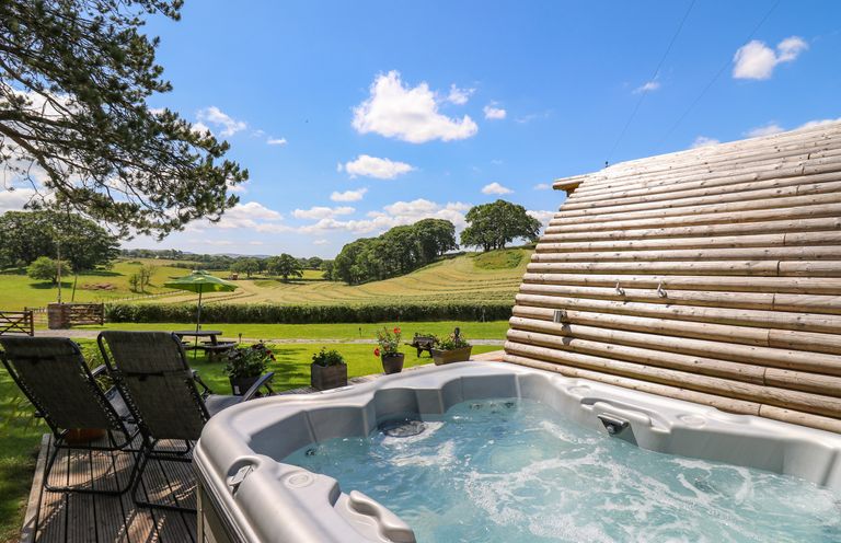 An outdoor hot tub beside a wooden log cabin with lounge chairs and a picnic table on a grassy area overlooking fields and trees at Pine Lodge in Llanddeusant near Llandovery