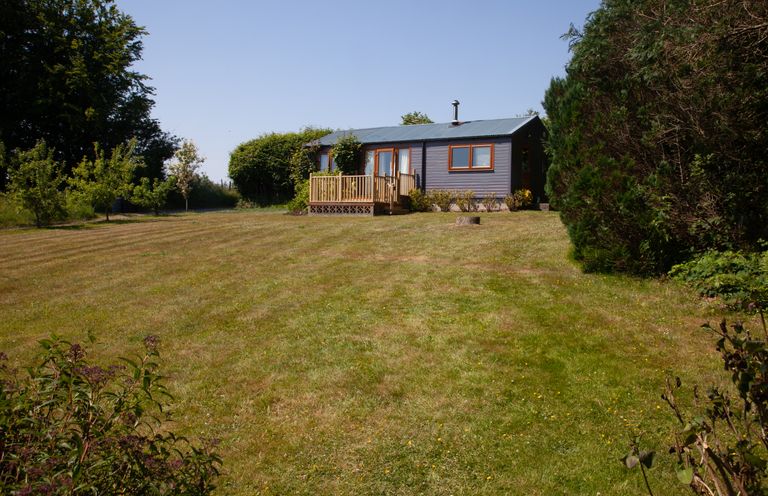 A small house with a wooden deck in a large garden with trees and bushes at Y Caban Glas in Rhydlewis near Brynhoffnant