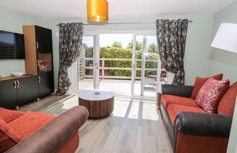 A living room with two sofas a round coffee table a black cabinet a wall mounted TV and sliding glass doors with floral curtains at Rose Lodge in High Newton-by-the-Sea near Embleton