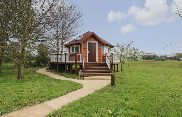 A small wooden cabin with a deck and steps surrounded by grass and trees at Honeybee Retreat in Pentney near Upper Marham