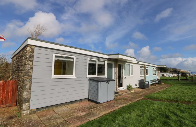 An exterior view of a single story building with windows a door a storage box and benches on a grassy area at Nanny's Little House in Sandown