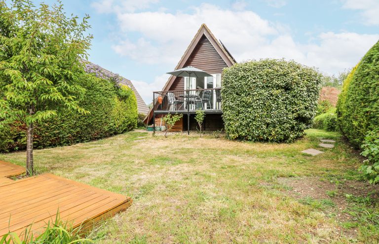 A garden with grass lawn and bushes with a wooden deck and an a frame house with a balcony and outdoor furniture at Kingfisher in Isleham