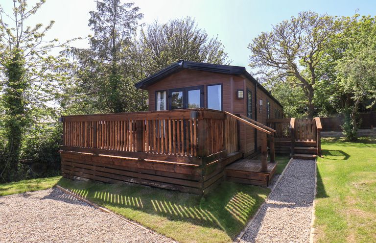 A wooden cabin with a deck and a gravel path surrounded by grass and trees at Buttercup Lodge in Runswick Bay near Staithes