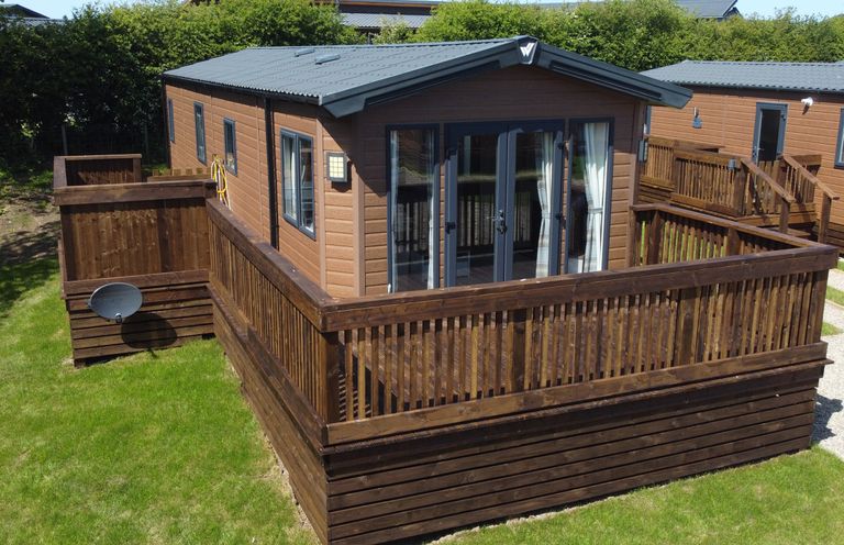 A wooden cabin with a fenced deck and glass doors on a grassy area at Rose Lodge in Runswick Bay near Staithes
