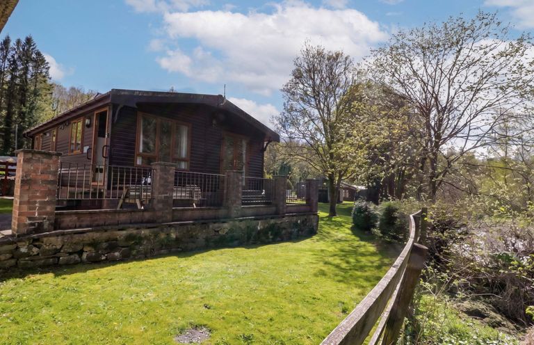 A lodge in a grassy area with trees and a fence at Elm Lodge in Keswick