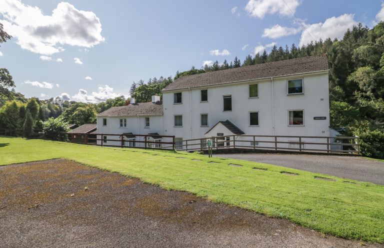 A white multi-story building with a sloped roof surrounded by grass and trees at Garden Cottage No 1 in Keswick