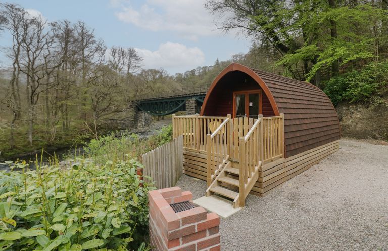 A wooden pod with a small deck and stairs near a river and a green metal bridge at Pod No 28 in Keswick