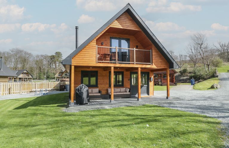 A wooden two-story house with a balcony and outdoor seating on the lawn at Sunset Lodge 5 in Llanbedr