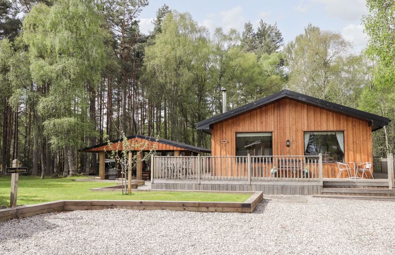 A wooden cabin with a deck and metal chairs surrounded by trees and a gravel area at Lovat Highland Bothy in Beauly