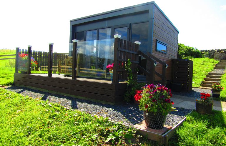 A small modern cabin with glass railing and flower pots in a grassy area at Balvicar Beag in Balvicar Isle Of Seil