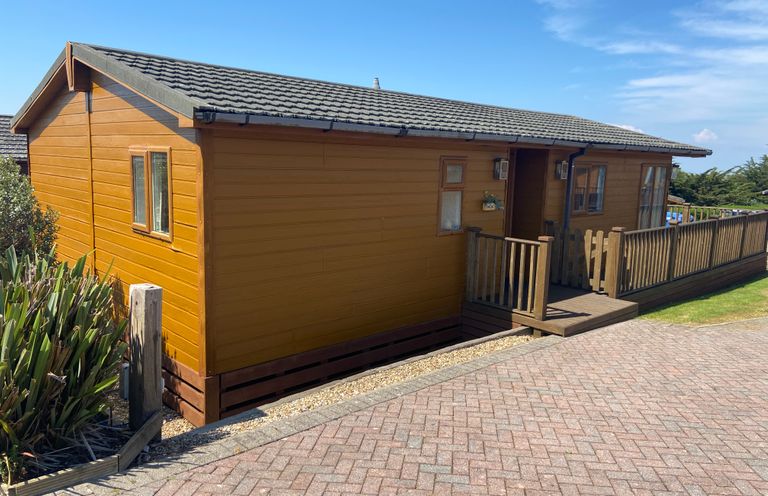 A wooden cabin with a small porch and railing next to a paved pathway at Mojo Mullacott near Ilfracombe