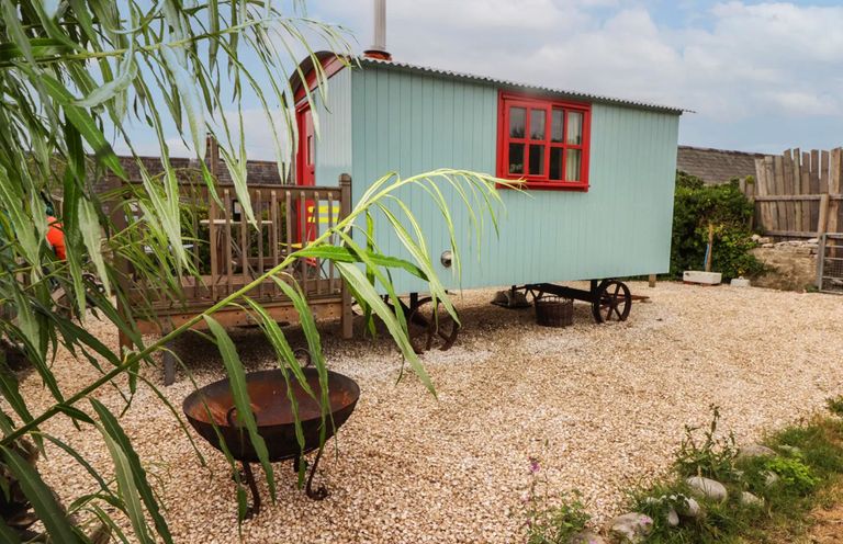 A small mobile hut with red windows and door on wheels next to a wooden deck with railing and a fire pit in a gravel yard at Shepherd's Hut in Southerndown near St Brides Major