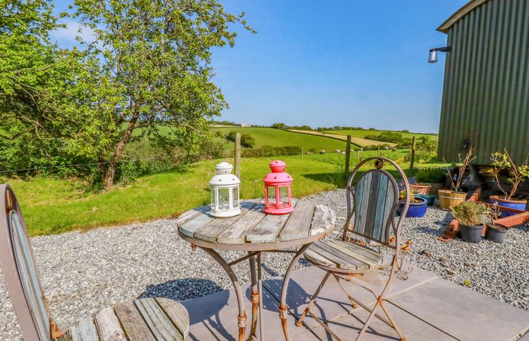 An outdoor patio with a round wooden table and two chairs with a tree and green fields in the background at Puddle Duck Shepherds Hut in Crackington Haven