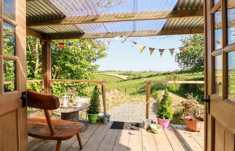 A wooden porch with chair and table surrounded by potted plants and bunting overlooking a rural landscape at Shepherds Retreat in Maxworthy near Crackington Haven