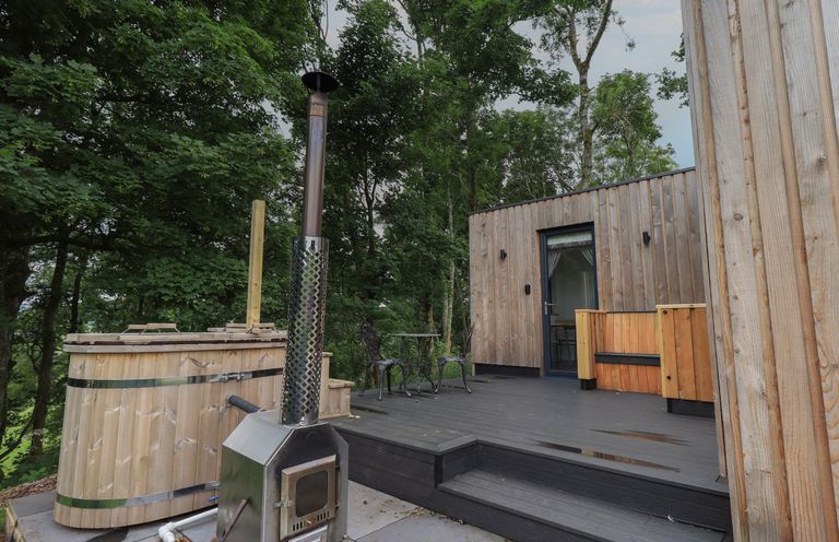 An outdoor deck with a wooden hot tub a metal stove a small table with two chairs and a wooden building at Yew Tree Cabin in Ullswater