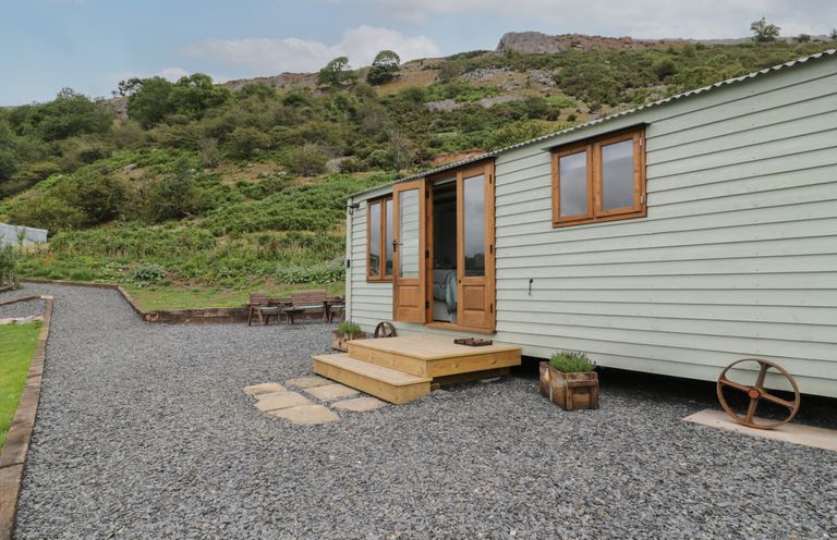The exterior of a light green wooden shepherds hut with open doors on a gravel surface with a hill in the background at Tan Y Castell Shepherds Hut in Llangollen