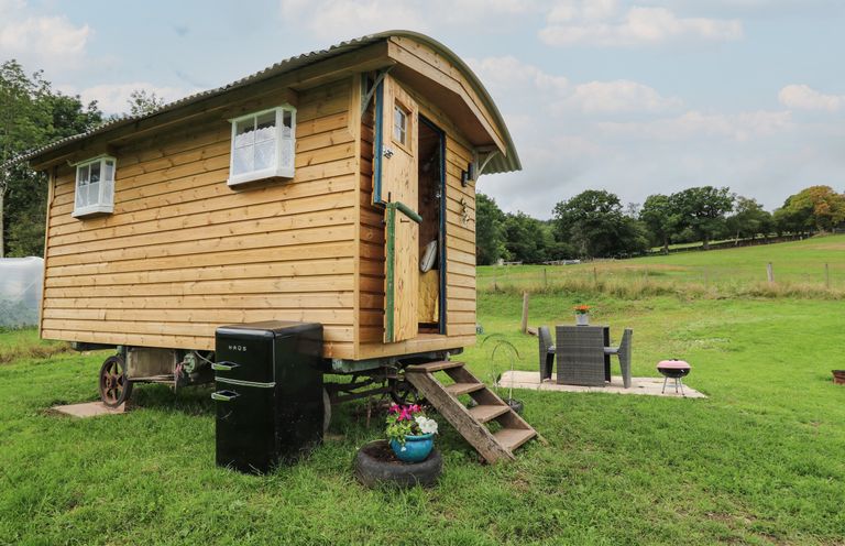 A wooden shepherd hut with small windows and steps leading to the doorway in a grassy field with a black refrigerator nearby and a patio set with chairs and a table at Ty Gwenyn in Talybont-On-Usk