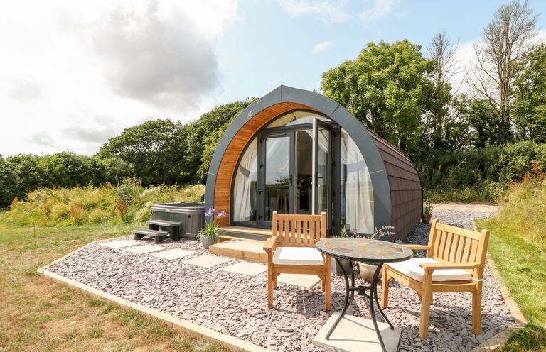 A small cabin with wooden furniture and a table outside on a gravel patio surrounded by grass and trees at Oak in Elmgate near Saltash