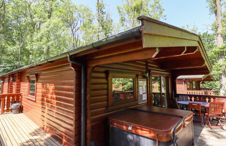 A wooden lodge with a covered porch featuring a hot tub and outdoor dining table surrounded by trees at 64 Acorn Lodge Kenwick Park in Louth
