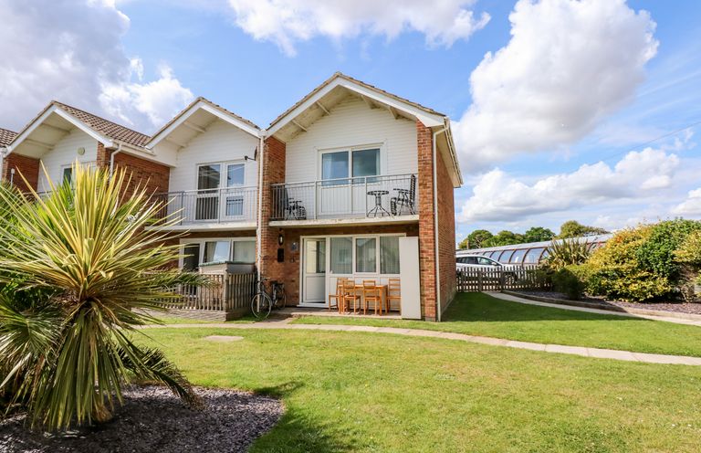 A two-story house with balconies and outdoor chairs on a lawn with a palm tree at 63 The Waterside Holiday Park in Corton