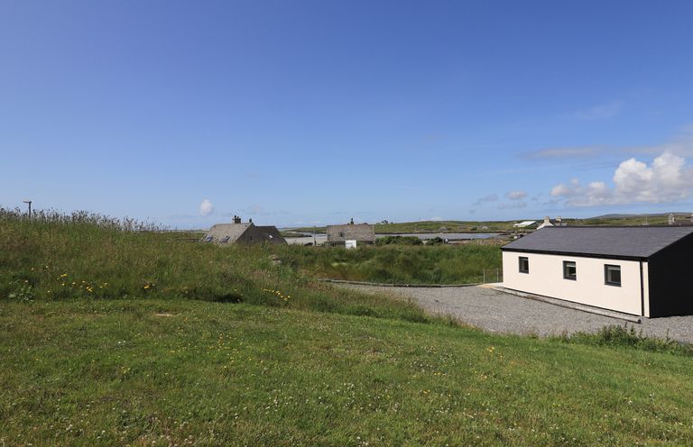 A grassy field with scattered houses and a gravel driveway under a blue sky at Eala Bhan Cottage in Clachan near Balivanich